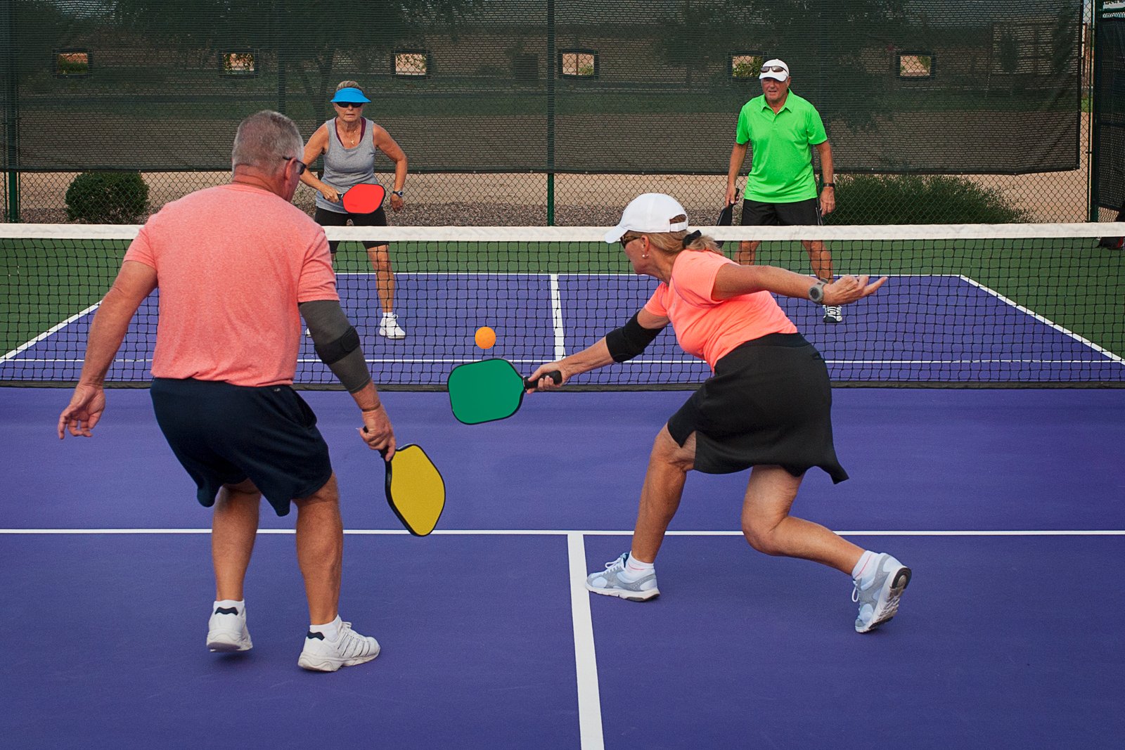 Four people playing pickleball on a purple court; a woman in a black skirt lunges to hit the yellow ball with a green paddle.